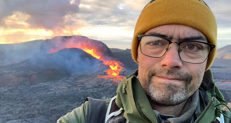 Han har mössa och glasögon. Glödande lava syns bakom honom.