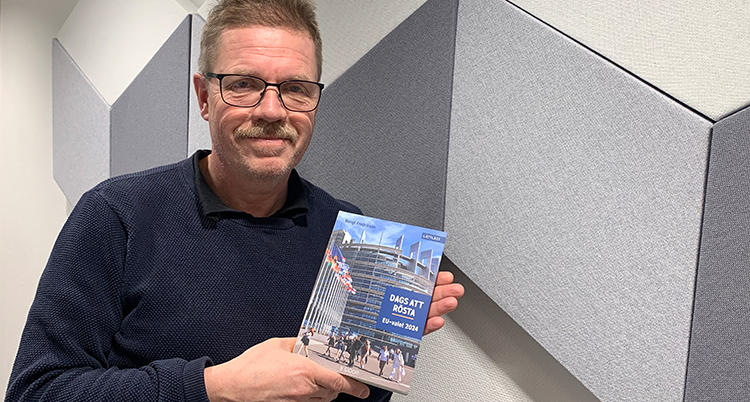 Han håller i boken. Han tittar in i kameran.