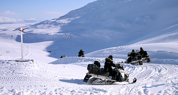 Det är en solig dag i fjällen. Några personer åker ut med snöskotrar.