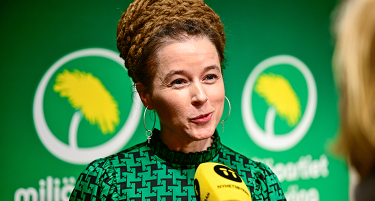 Hon pratar i en mikrofon. Hon har grön klänning och står framför en grön vägg med Miljöpartiets logga på.