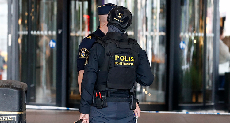 Två poliser står utanför en entré med ryggen mot fotografen. Det står Bombtekniker på en av dem.