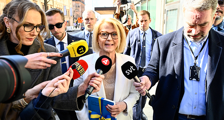 Hon håller i en bunt med papper. Många journalister står runt henne med mikrofoner.