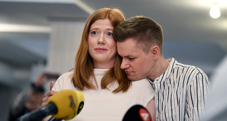 Han kramar om henne. De står framför flera mikrofoner och pratar.