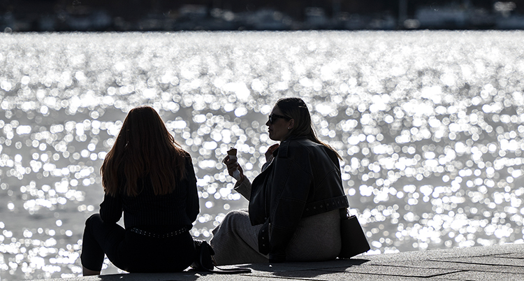 Två personer sitter i solen vid vattnet.