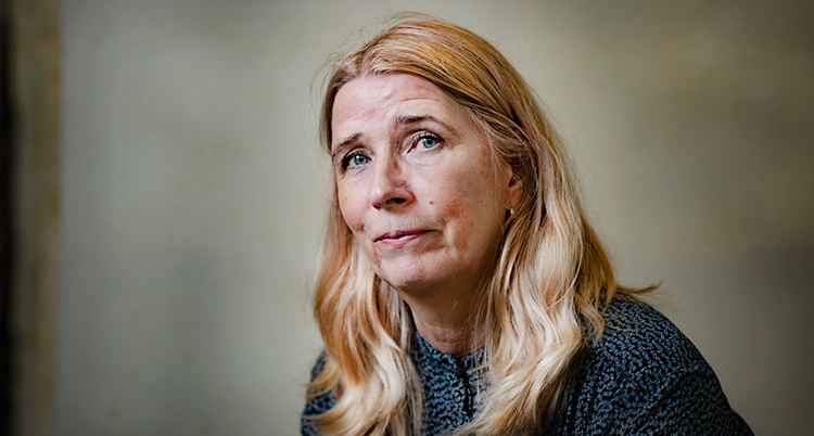 Hon tittar in i kameran, Hon har hår till axlarna och en blus på sig.