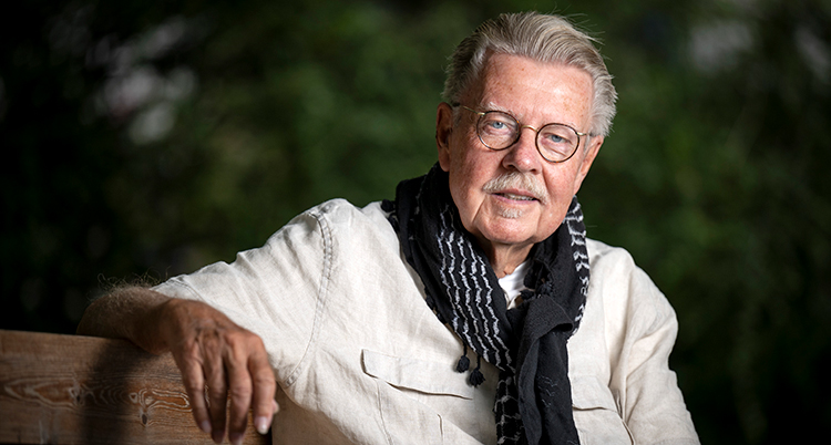 Han sitter på en bänk och tittar in i kameran. Bakom syns gröna buskar.