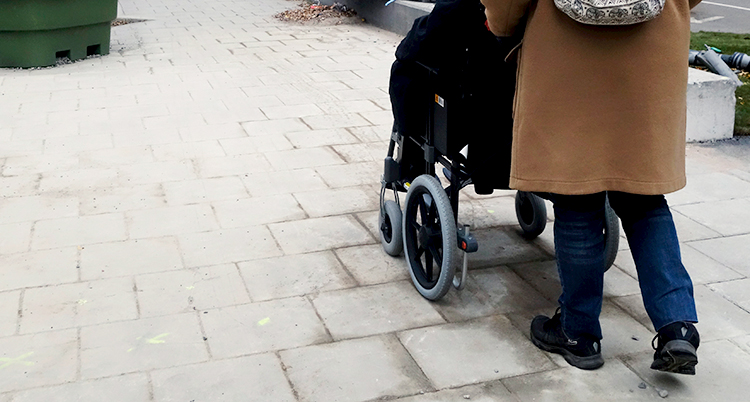 En person sitter i en rullstol. Personen blir körd av en annan person på en trottoar.