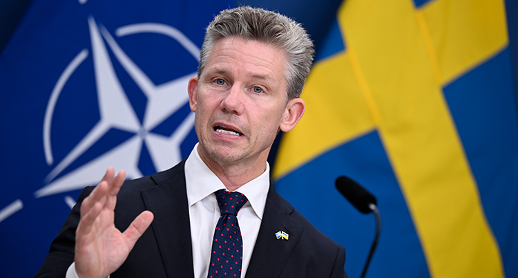 Han har slips och kavaj. Han pratar framför en mikrofon.