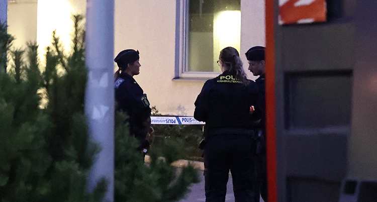 Tre poliser står vid ett hus med lägenheter. Ett polisband syns bakom dem.