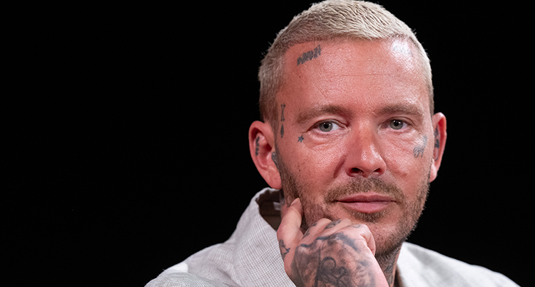 Han tittar in i kameran. Han tar tatueringar i ansiktet.