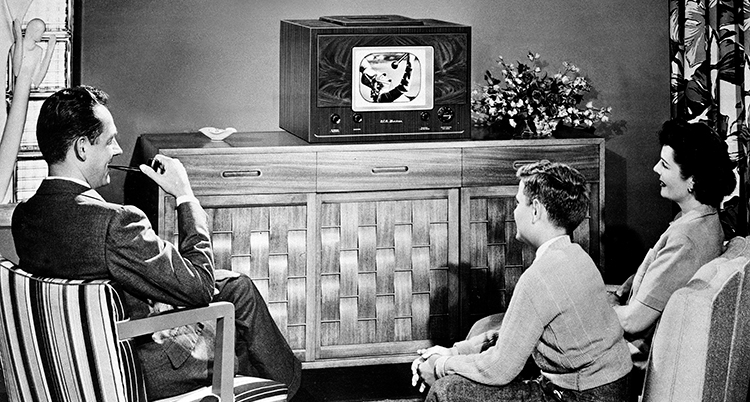De sitter i ett vardagsrum och tittar på en liten tv. Mannen röker pipa.