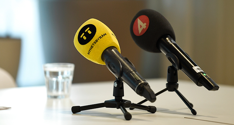 Två mikrofoner står på ett bord. Det är journalisters mikrofoner. På en av dem står det TT Nyhetsbyrån. På den andra står det TV4.