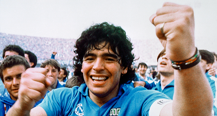 På en fotbollsplan. Maradona är glad och knyter sina nävar. Massa människor runt honom.