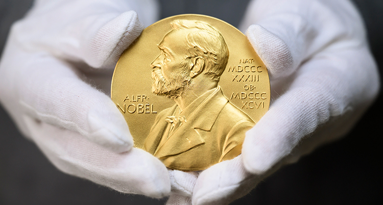 En person med vita handskar håller fram en guldig medalj med en bild av Alfred Nobel.