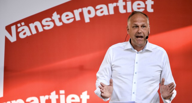 Vänsterpartiets ledare Jonas Sjöstedt i vit skjorta framför ett rött skynke som det står Vänsterpartiet på.