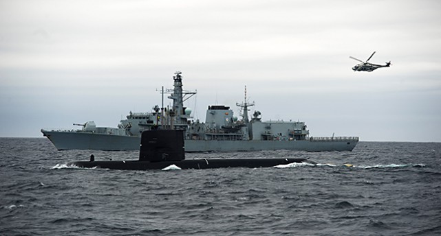 En ubåt på ett hav. I bakgrunden ett militärt fartyg.