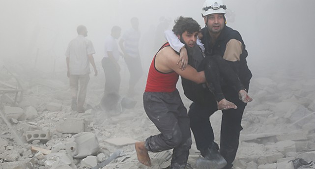 Två människor bär en tredje person från ett bombat område. Bakom dem finns rök, stenar och grus. I röken syns andra människor.