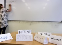 En kateder med namnlappar på elever i förgrunden. Bakom katedern ser man en lärare som skriver på en whiteboard.