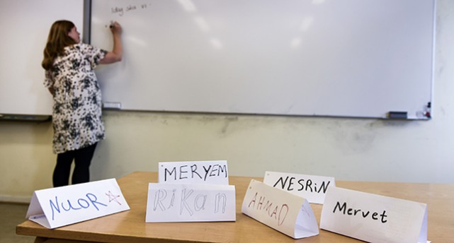 En kateder med namnlappar på elever i förgrunden. Bakom katedern ser man en lärare som skriver på en whiteboard.