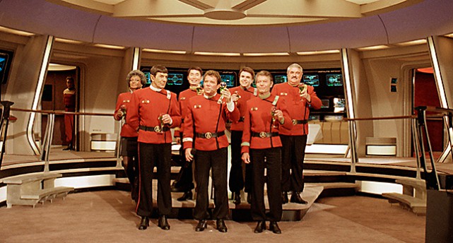Flera av medlemmarna i Star Trek. De står upp i sin farkost. Alla har röda jackor med svarta skärp och svarta byxor.