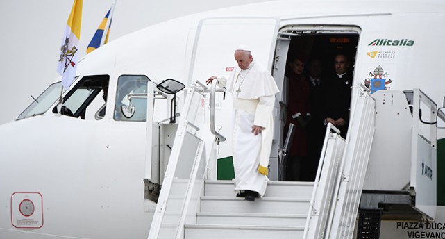 Påven klädd i vit kaftan och vit kalott och svarta skor kliver ner för flygplanstrappan som är vit. Han håller upp kjolen lite med ena handen. I bakgrunden syns det vita flygplanet.