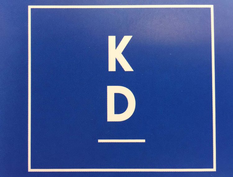 En blå ruta med en tunn vit ram. Bokstaven K ovanför bokstaven D och ett tunnt vitt streck under.