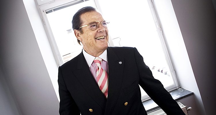 Roger Moore står framför ett fönster och ler