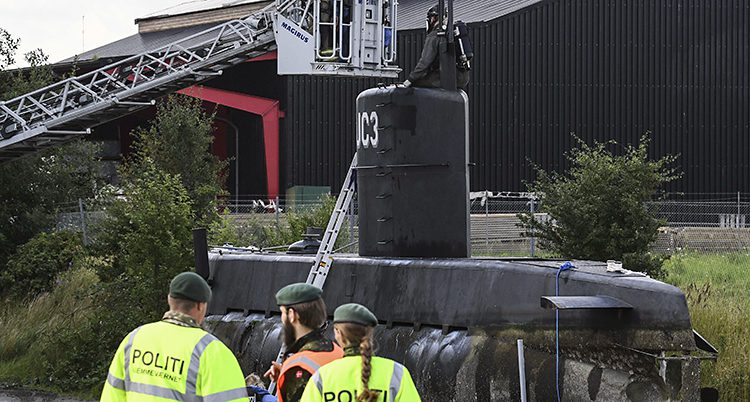 Poliser i gula reflexjackor står nedanför ubåten som står på marken. En stege står lutad mot ubåten. En svartklädd person med mask för ansiktet sitter högst upp på ubåten.