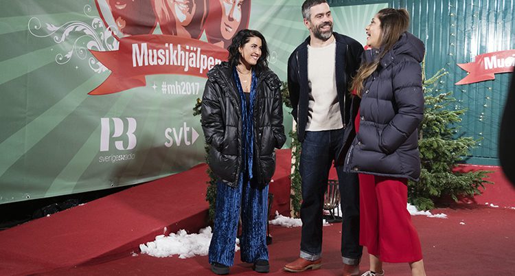 Molly Sandén, Kalle Zackari Wahlström och Farah Abadi står framför en stor skylt med Musikhjälpens logga.
