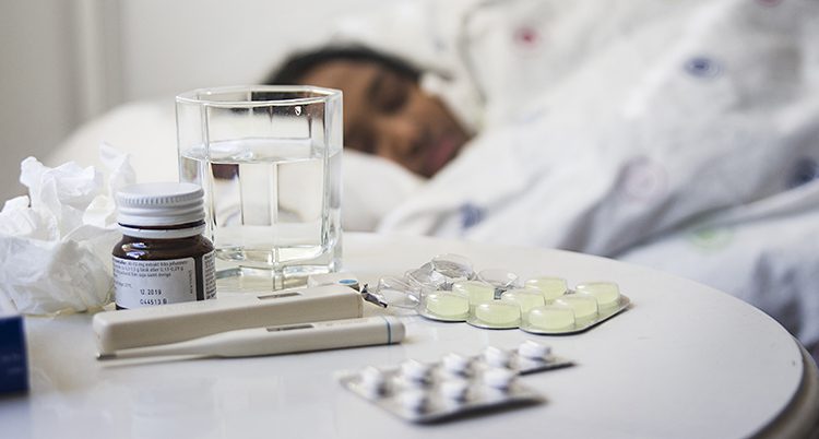 En person ligger i en säng med tabletter, vattenglas och näsdukar framför sig.