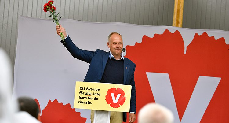 Jonas Sjöstedt vid talarstolen. Han vinkar med en bukett röda blommor i handen.