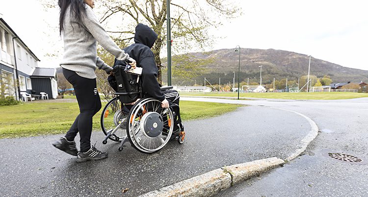 En gata utomhus. En person sitter i en rullstol. En annan person kör rullstolen. Vi ser dem bakifrån