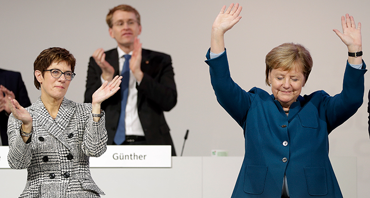 Merkel lyfter händerna och vinkar till publiken. Annegret Kramp-Karrenbauer står bredvid och applåderar.