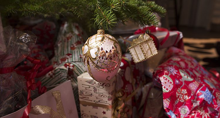 En bild på en del av en julgran. Vi ser en gren med en rosa förgylld julgranskula och några inslagna julklappar under.