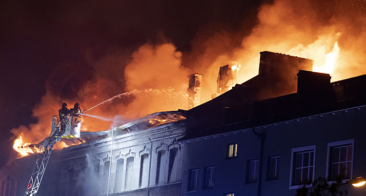 Brandmän i en korg högt uppe på en stegensprutar vatte över taget. Det brinner i taker. Det är mörkt och lågorna är orangea.