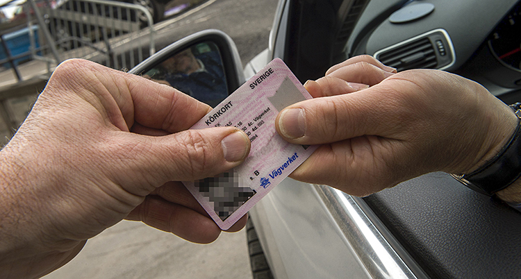 En person sitter i en bil och sträcker ut ett körkort. En annan hand tar emot körkortet.