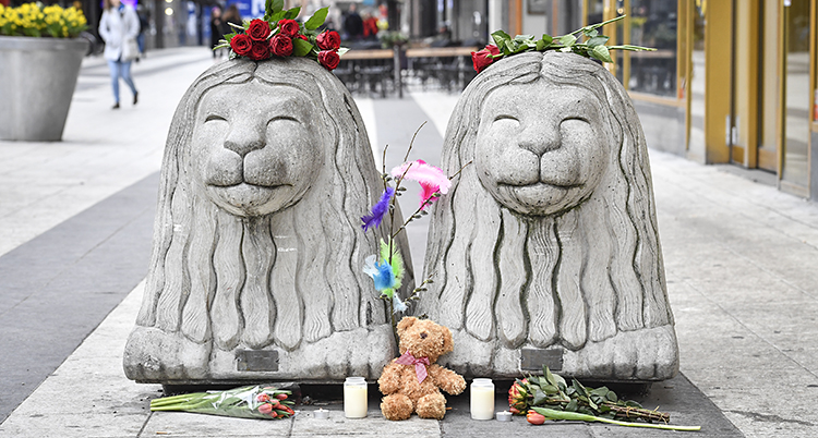 Två stora lejon i betong. Folk har lagt blommor bredvid och på lejonen.