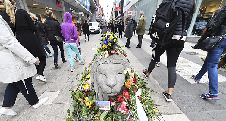 Människor går förbi ett lejon i betong. Det ligger en massa blommor runt lejonet.