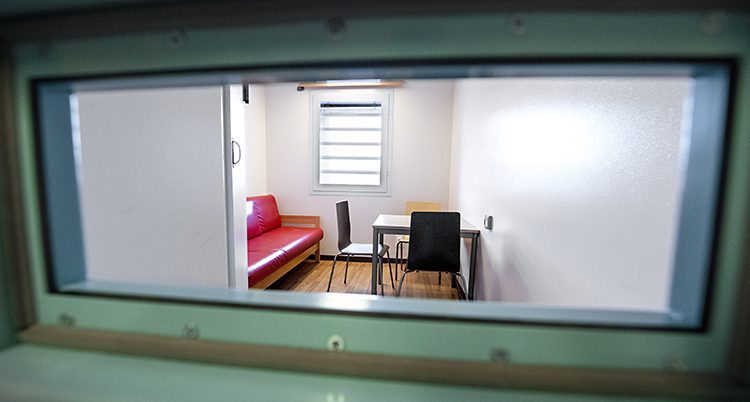 Bilden är tagen genom en liten ruta i en dörr. På andra sidan är en cell på ett häkte. Där syns bland en stol och ett bord.