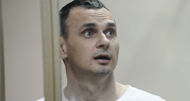 Oleg Sentsov sitter bakom galler i en rättegångssal. Han har en vit tröja på sig. Han spärrar upp ögonen och tittar på något utanför bilden.