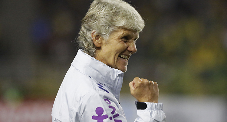 Pia Sundhage har kort, grått hår. Hon har på sig en vit träningsjacka med lila detaljer. Hon ler och knyter näven i en segergest.