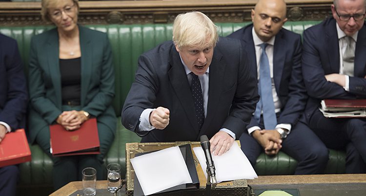 Boris Johnson står i riksdagen och pratar. Han gör en rörelse med armen och ser engagerad ut. På bänken bakom honom sitter flera andra politiker.