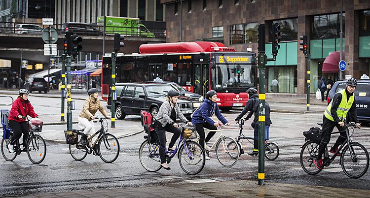 Flera personer cyklar i stadstrafik. En röd buss syns i bakgrunden.