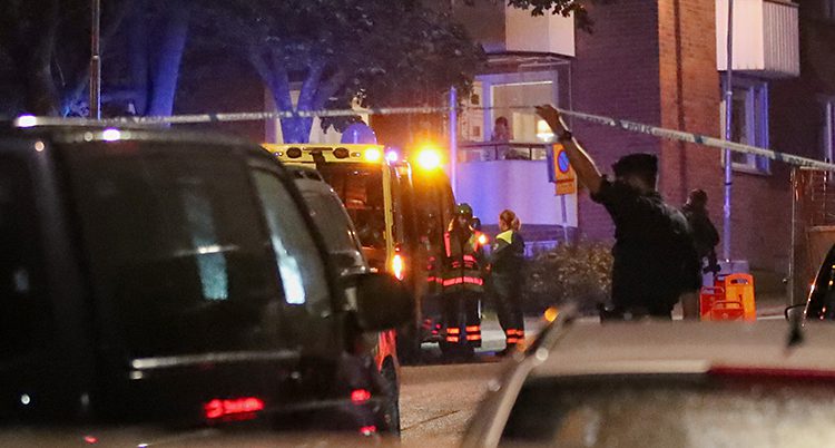 En polis vid en bil lyfter på ett band som spärrar av. I bakgrunden står två personer vid en ambulans. En kvinna tittar ut från en balkong.