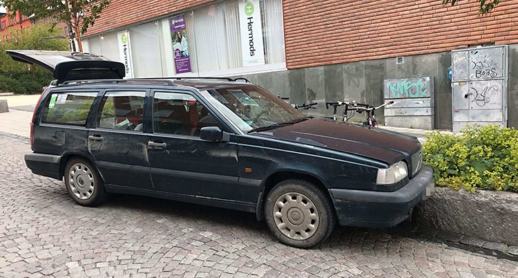 En mörk bil står på en gata med kullerstenar. Det är en mörk Volvo kombi. Bakluckan är öppen. Det är i stadsmiljö, med en liten plantering och ett hus bakom.