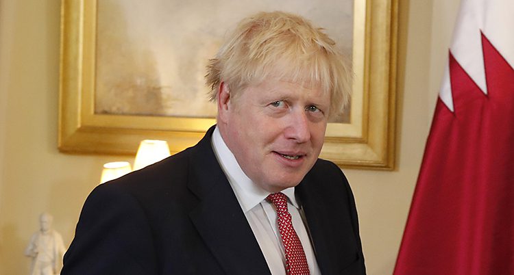 Storbritanniens ledare Boris Johnson står i ett rum. I bakgrunden syns en tavla och en röd och vit flagga. Boris Johnson har svart kavaj, vit skjorta och röd slips. Hans hår är blont och spretigt.