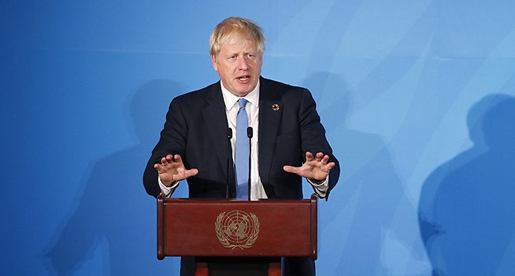 Boris Johnson står och håller ett tal. Han står vid en röd pulpet med FN:s symbol på och pratar i en mikrofon. Han har en mörk kostym, vit skjorta och blå slips. Väggen bakom är blå.