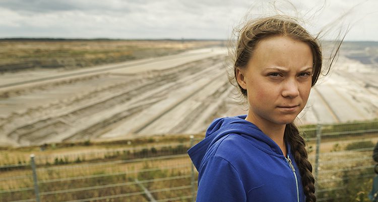 Greta Thunberg tittar in i kameran. Hennes hår blåser lite i vinden. Hon har en fläta och en blå tröja. I bakgrunden syns ett stort fält.
