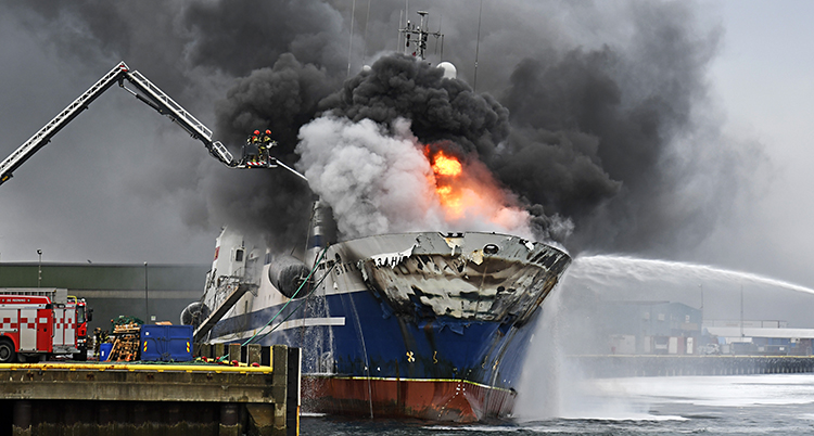 På bilden syns en stor båt som ligger i en hamn. Det syns eld på båten och det kommer en massa rök.