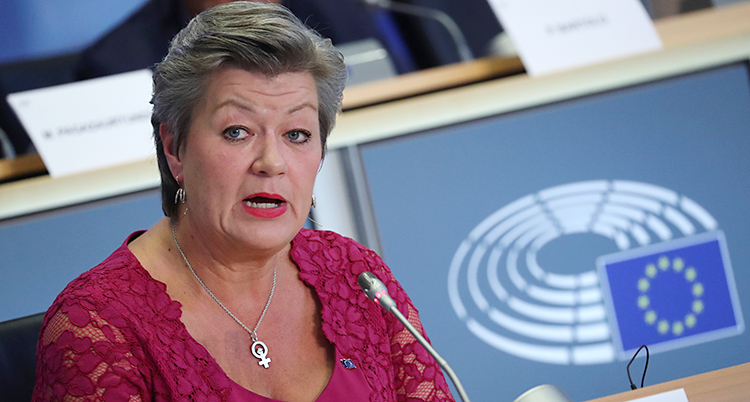 Ylva Johansson har röd spetströja och sitter vid ett bord med mikrofon